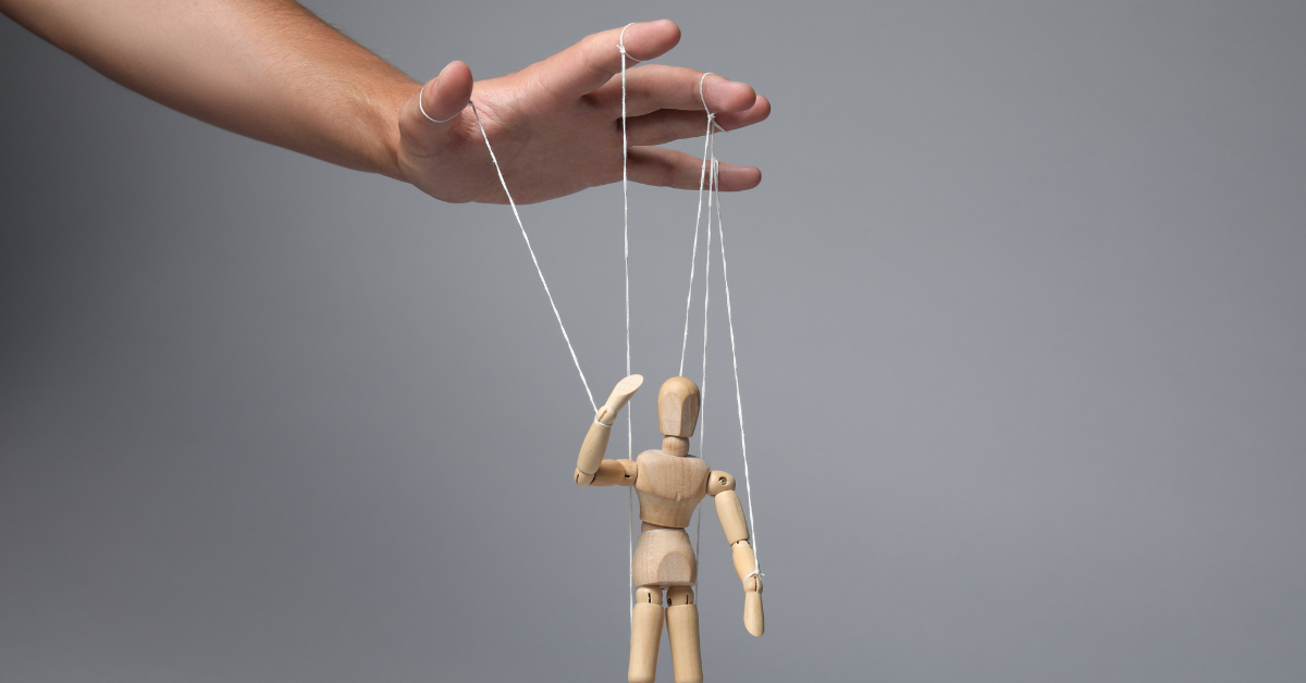 Drevená bábka, ktorú motúzikmi manipuluje ruka, symbolizujúca kontrolu a manipuláciu.